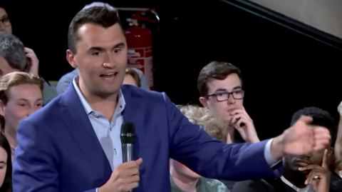 Charlie Kirk in a blue suit and shirt, holding a microphone, with short dark brown hair, gesticulating and talking into a microphone. There are people sitting behind him spectating.