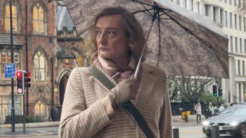 Julian Johnson-Munday stands outside the court under a spotted umbrella. He has shoulder-length wavy hair and is wearing a scarf, a beige checked jacket and beige gloves.