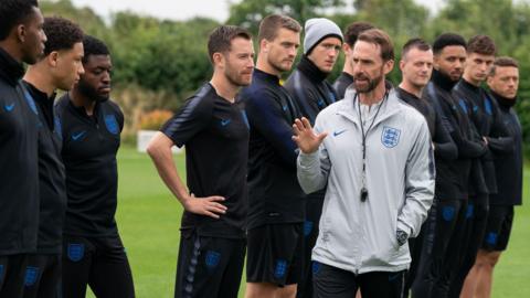 A man with a beard and short, brown hair in a grey sweatshirt with England's three lions on it ,and a whistle around his neck is talking to a line of men dressed in black football kit.