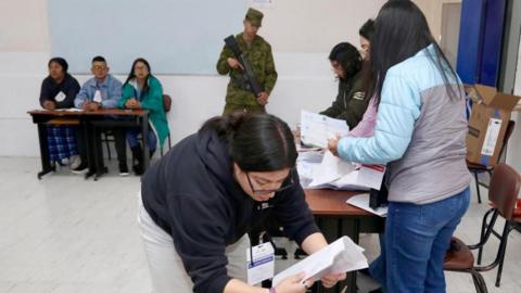 A group of women count ballots in Ecuador as an armed soldier keeps guard behind them