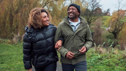 A father and daughter walk arm in arm through the countryside. They are both wearing warm coats, and the father is a wearing a black woolly hat. Both are smiling as they walk.