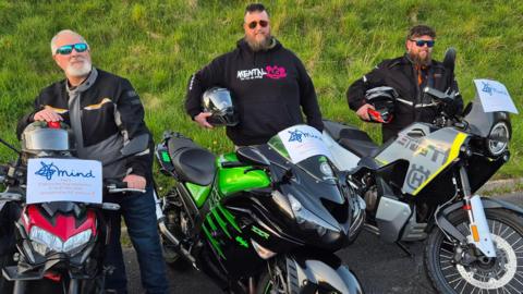 Christopher Smith is pictured with his father and brother in law. All three are wearing biker gear and holding their helmets under their arms. They are smiling at the camera while standing next to their motorbikes. Each motorbike has a small sign on the front that says Mind with a little blurb about their fundraiser.