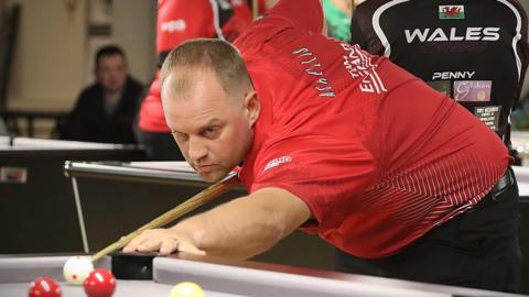 A man bent over a pool table with his left hand on the edge of the table and a wooden pool cue in his right hand. He is aiming for a white ball, and there are two red balls and a yellow ball near him. He is wearing a red T-shirt with white writing on it and black trousers. He has short brown hair. There are more pool tables and people in the background.