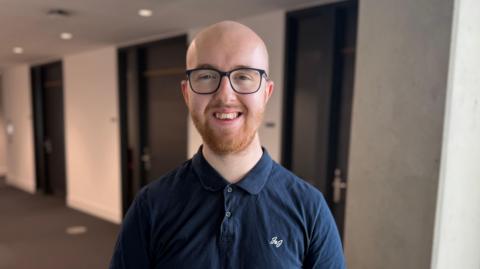 A young man standing in a hallway. He is wearing a blue polo shirt, and reading glasses.
