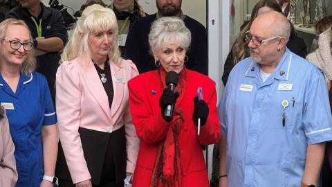 Anita Dobson looking at the camera wearing a red coat and scarf with black gloves. To her left is Julie Rowley, Director of retail at Thames Hospice she is wearing a black dress and pink short jacket.