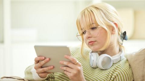 Side view of smiling female teenage GCSE student sitting on sofa in the living room at home, holding tablet in hands, revising with flashcards.