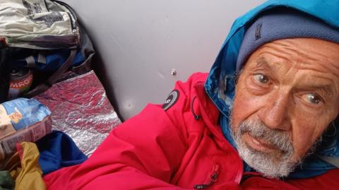 Selfie of Nick Brooke wearing a red coat and another blue waterproof jacket underneath with the hood up. He appears to be laying on the floor and leaning on a wall, surrounded by his belongings, including a rucksack. He has a tanned face and a full grey beard.