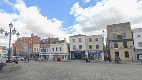 A row of shops in Boston town centre. People can be seen walking on the paths. The sky is blue with large white clouds. 