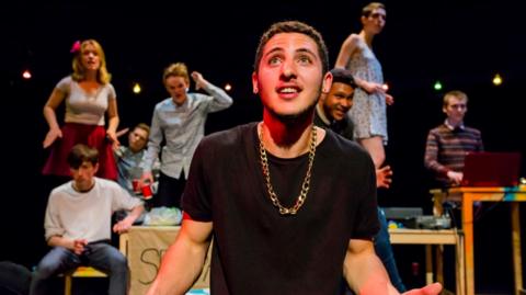 A young male theatre performer in a black T-shirt and wearing a large gold chain looks up with his arms outspread. Behind him, on stage at Bristol Old Vic, are other young performers - not in focus - in various poses
