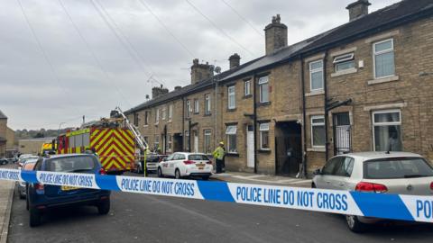 'Police line do not cross' tape with a fire engine on a residential street with cars parked there too