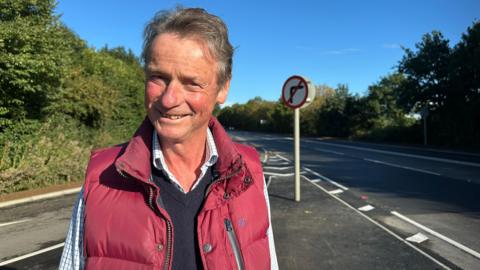 Gideon Sumption. He has short grey hair and is wearing a checked shirt, a navy blue jumper and a red gilet. He is pictured near a road junction. He is looking at the camera and smiling.
