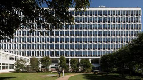 A large mid-rise building of white concrete and blue windows.The building facade is seen from a grass courtyard that also features multiple short trees.