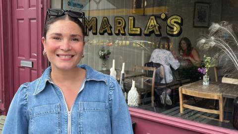 A person with dark hair and wearing a blue denim dress stands outside a maroon-coloured cafe. The window has the word "MARLA'S" written across it in large golden letters. The interior of the building can be seen with people sitting at tables and several vases of flowers. Benches are also visible in the background.