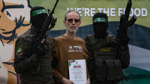 Ohad Ben-Ami in a brown sweatshirt, standing between two Hamas fighters holding guns and with black balaclavas covering their faces. 