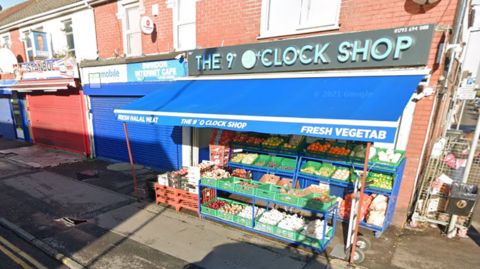 A shop front with a dark grey sign with blue letters that read The 9 O'Clock Shop. A blue store covering is over boxes of fruit and vegetables facing the pavement with a couple of other businesses to the left.