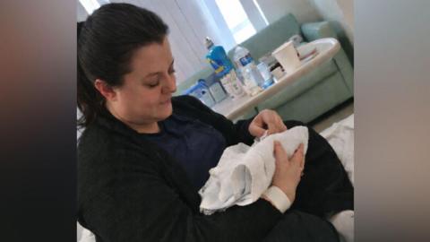 Ashley Waugh sitting on a hospital bed with Benjamin in her arms wrapped in a blanket. She has brown hair which is tied back in a ponytail and she is looking down at the baby with her legs crossed beneath her. In the room behind her there is a table with various empty bottled drinks and a blue sofa. She is wearing all black and has a hospital bracelet around her wrist.