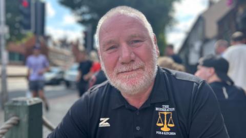 Andy Becalick is wearing a black T-shirt with The Moderation sewn into it. He has short, white hair and has a white beard. He is sat outside and people are walking past, presumably on the way to the festival.