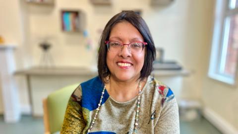Andrea Thasan looks directly at the camera. She is wearing red-metal trimmed glasses and is wearing red lipstick. She has black hair cut into a bob swept to the side. Thasan is wearing a long-sleeved multi-coloured top and has a string of beads on. She is smiling. Thasan is sitting on a green chair in a training room with a fireplace and a cream wall. The background is slightly blurred.