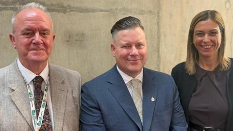 (Left to right) A man with a grey suit, white shirt and tie with a North Northamptonshire Council-branded white and blue lanyard round his neck. A man in the middle with a blue jacket, white shirt and tie wearing a pin badge. A woman with a black jacket and brown dress with belt. All three are looking directly at the camera.