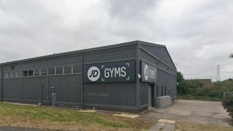 The side of a grey building with corrugated metal sides and a large sign which reads JD Gyms