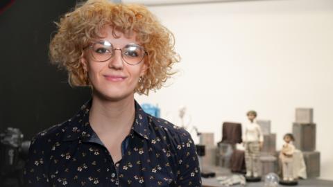 Laura Tofarides. Mae ganddi wallt golau a chyrliog ac mae'n gwisgo sbectol a chrys blodeuog.