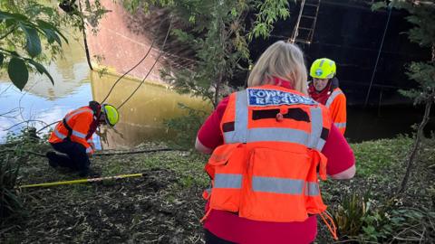 People with bright orange high-visibility jackets at a riverbank