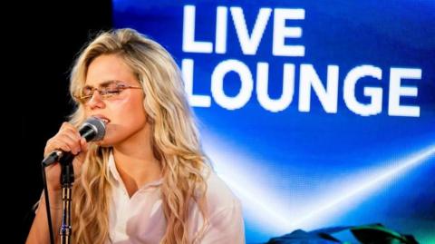 Artist and singer Reneé Rapp signing into a mic with the Radio 1 Live Lounge logo in the background