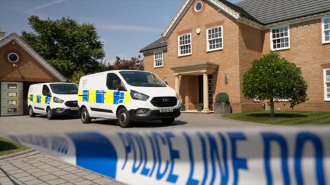 Two police forensic vans are on the drive in front of a large brick house. The front door has a veranda with two stone pillars. White and blue police tape that says 'Police line do not cross' is in the foreground.
