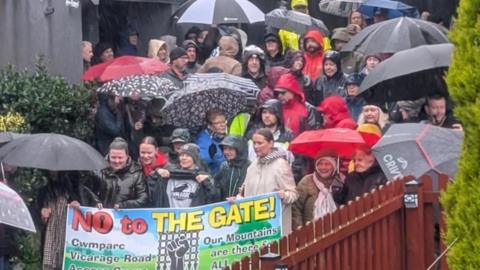 Picture of around 40 people gathered outside a home smiling towards a camera. Most can be seen stood in the rain wearing rain coats and carry red, black and grey umbrellas. 