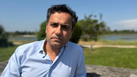 Former MP Rehman Chishti, with dark curly hair and a light blue shirt, sitting on a bench in front of trees and water