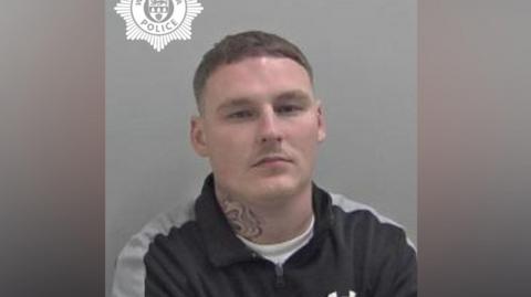 A mugshot of a man with brown hair. He is wearing a black and grey top and has a tattoo on his neck.