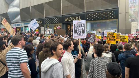 Crowds of people stand outside a row of shops, with several people holding up protest signs such as 'Jobs & 91ȱs Not Racism'