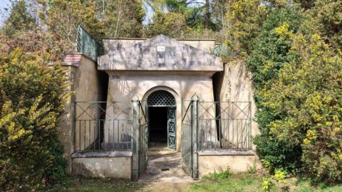 The entrance to a mausoleum. There is a green gate at the front, with plants surrounding the entrance.