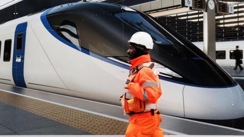 A construction worker in fluorescent orange clothing and white hard hat walks past an advert for HS2