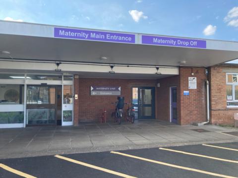 A single-storey red brick hospital maternity unit, with signs above doors saying "maternity main entrance" and "maternity drop off". Yellow lines of parking bays are on the tarmac in front of the building. 