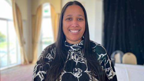 Claire Hutton is standing in a conference room with cream curtains. She has straight long black hair and is wearing a black top with white flowers and smiling.