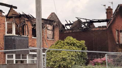 Two houses, with collapsed roofs, can be seen behind metal fencing. There are gardens in the foreground and a telegraph pole.