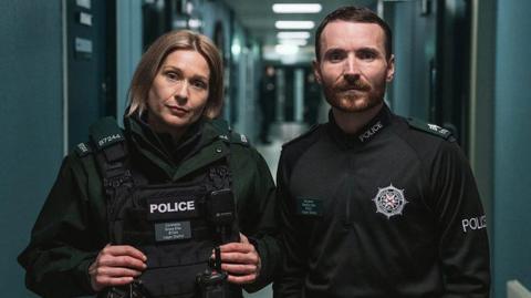 Sian Brooke and Martin McCann stand as their characters Grace Ellis and Stevie Neill in the corridor of a police station looking at the camera. They are wearing police uniforms, with Sian Brooke on the left wearing a large black vest with her name and station code. 