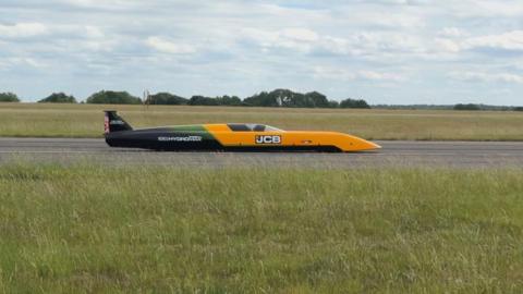 A photograph of the JCB vehicle on a runway. It is a long vehicle with a yellow front with "JCB" on it. The back half of the car is dark green and black with white writing. To the front and back is grass and it is on a grey surface. 