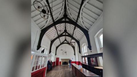 The empty waiting room is long and narrow. It has a bar on the right of the photo and large, almost floor-to-ceiling windows on either side. The walls are painted white, with some red features. The high vaulted ceiling is painted white with some arches painted in a dark colour as a feature. There is a large fireplace at the end of the room and three people are standing in the empty space.