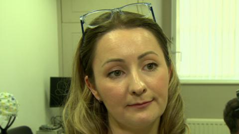 Dr Kathy Ruddy is wearing a white shirt and has long blonde hair. Her glasses are on top of her head and she has a silver pendant necklace on.