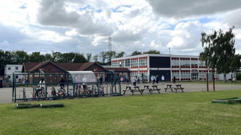 Hazlewood Primary School. There is a span of grass, then a concrete playground with a bike rack and picnic tables in front of the school buildings themselves. Two large buildings are visible. One, which appears to be older, is a single-storey building with a slanted roof. The other is a modern two-storey building with rows of glass windows on every side and a flat roof. Parents wait outside for their children to finish school.