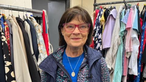 Mary Small wearing blue top, jacket and red glasses, with rack of clothes either side in the background