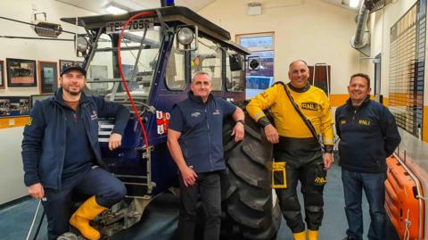 Four lifeboatmen in RNLI outfits with a wheeled vehicle inside a building.