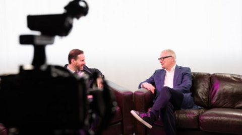 Mayor Richard Parker is sat crossed legged on a brown leather sofa in a television studio that is brightly lit and has white curtains draped in the back. He is sat opposite a man with slicked back brown hair. Mr Parker has grey short hair, wears black framed glasses, a white shirt underneath a blue blazer jacket.