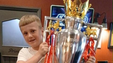 Cole, 11, with blond hair wearing a white t-shirt holds up the Premier League trophy in a pub. He is smiling.