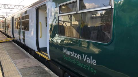 A green and white train stopped at a station with its door open. There is a ramp to one of the doors to the left and several windows on the train. 