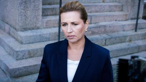 Denmark's Prime Minister Mette Frederiksen stands in front of stone steps wearing a dark blue suit jacket. Her hair is pulled back and she has a serious expression