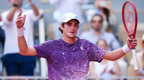 Joao Fonseca, wearing a purple t-shirt flecked with white and a white cap, holds his arms aloft with his red racquet in his left hand
