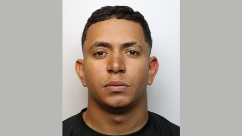 A mugshot of a man looking at the camera with a serious expression. He has short black hair and is wearing a black top